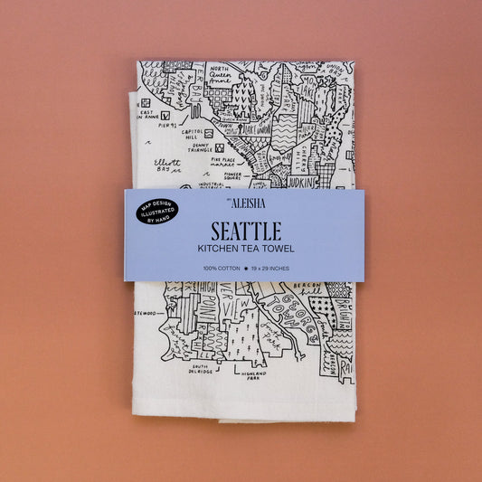 A folded white cotton kitchen tea towel with a black hand-drawn map of Seattle, wrapped with a light blue paper band that reads 'ALEHIA SEATTLE KITCHEN TEA TOWEL', set against a solid terracotta background.