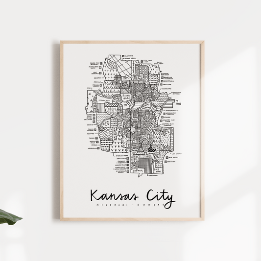A framed print of a Kansas City neighborhood map with the text 'KANSAS CITY' on the map, displayed above a green potted plant. The map is in black and white, showing detailed city boundaries and labels.