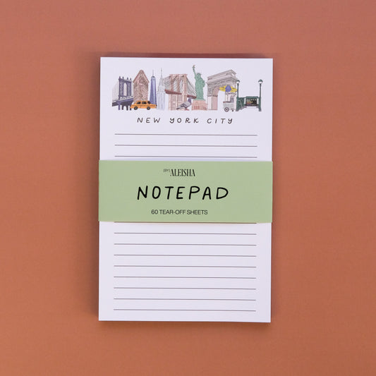 A small notepad with New York City skyline illustrations at the top, held by a light green paper band reading 'ALESHA NOTEPAD' on a solid terracotta background.