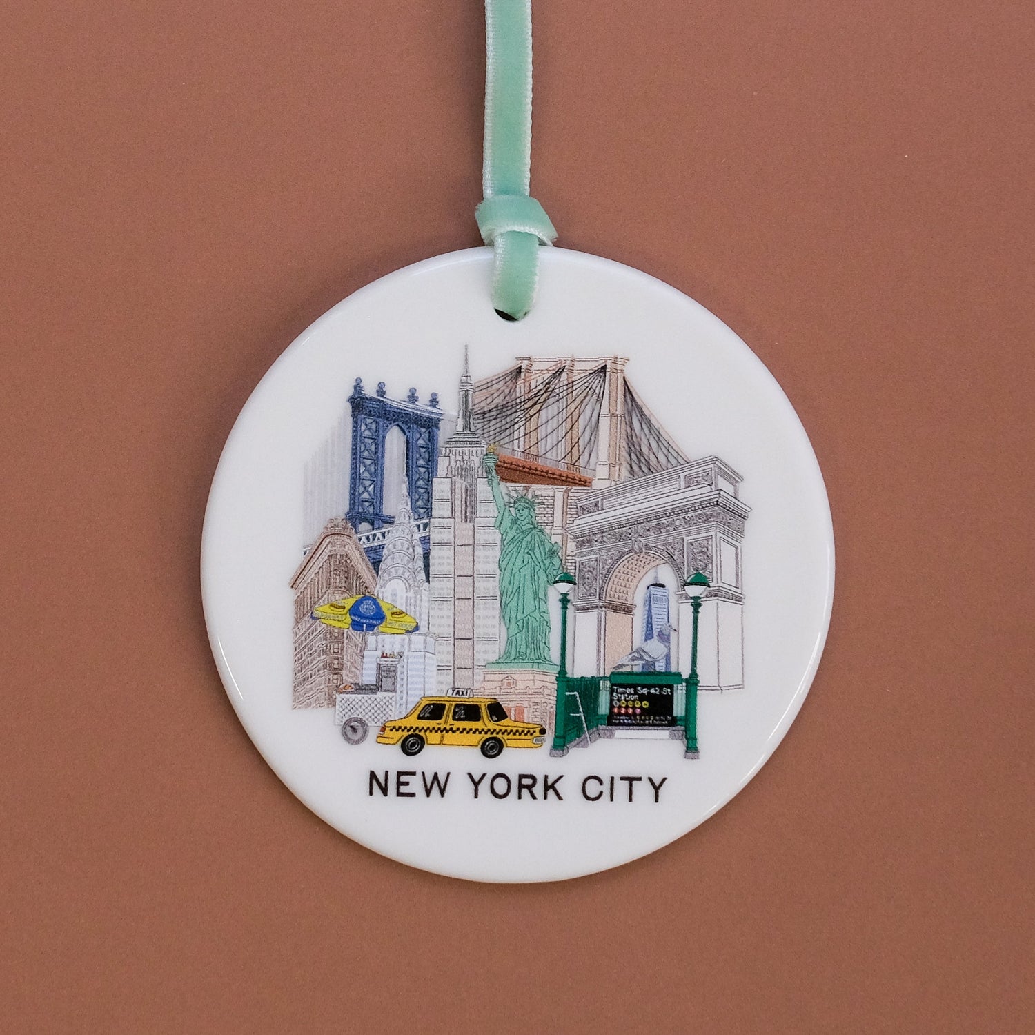 A round white ceramic ornament featuring an illustrated collage of New York City landmarks, including the Statue of Liberty, Brooklyn Bridge, and a yellow taxi, with the text 'NEW YORK CITY' at the bottom. The ornament hangs from a mint green ribbon against a solid terracotta brown background.