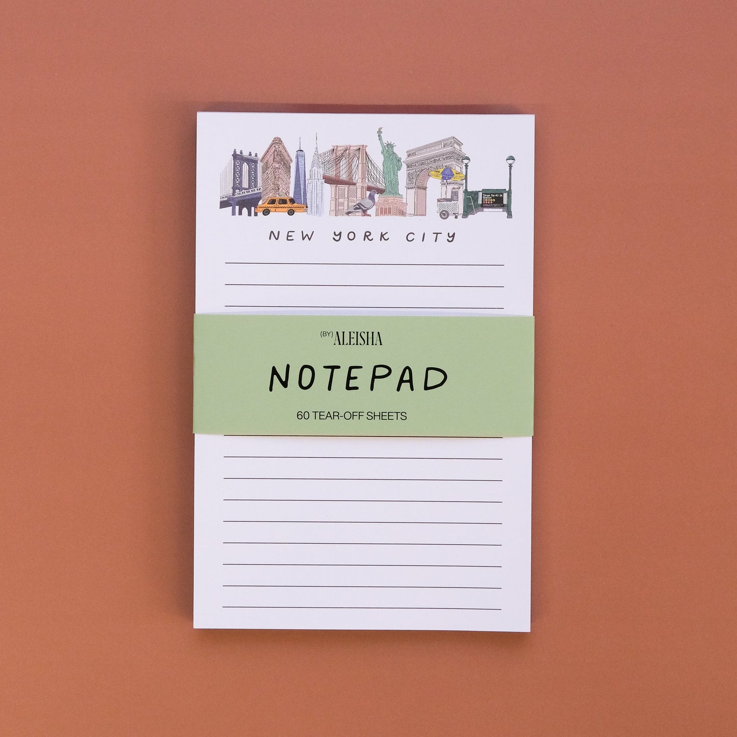 A small notepad with New York City skyline illustrations at the top, held by a light green paper band reading 'ALESHA NOTEPAD' on a solid terracotta background.