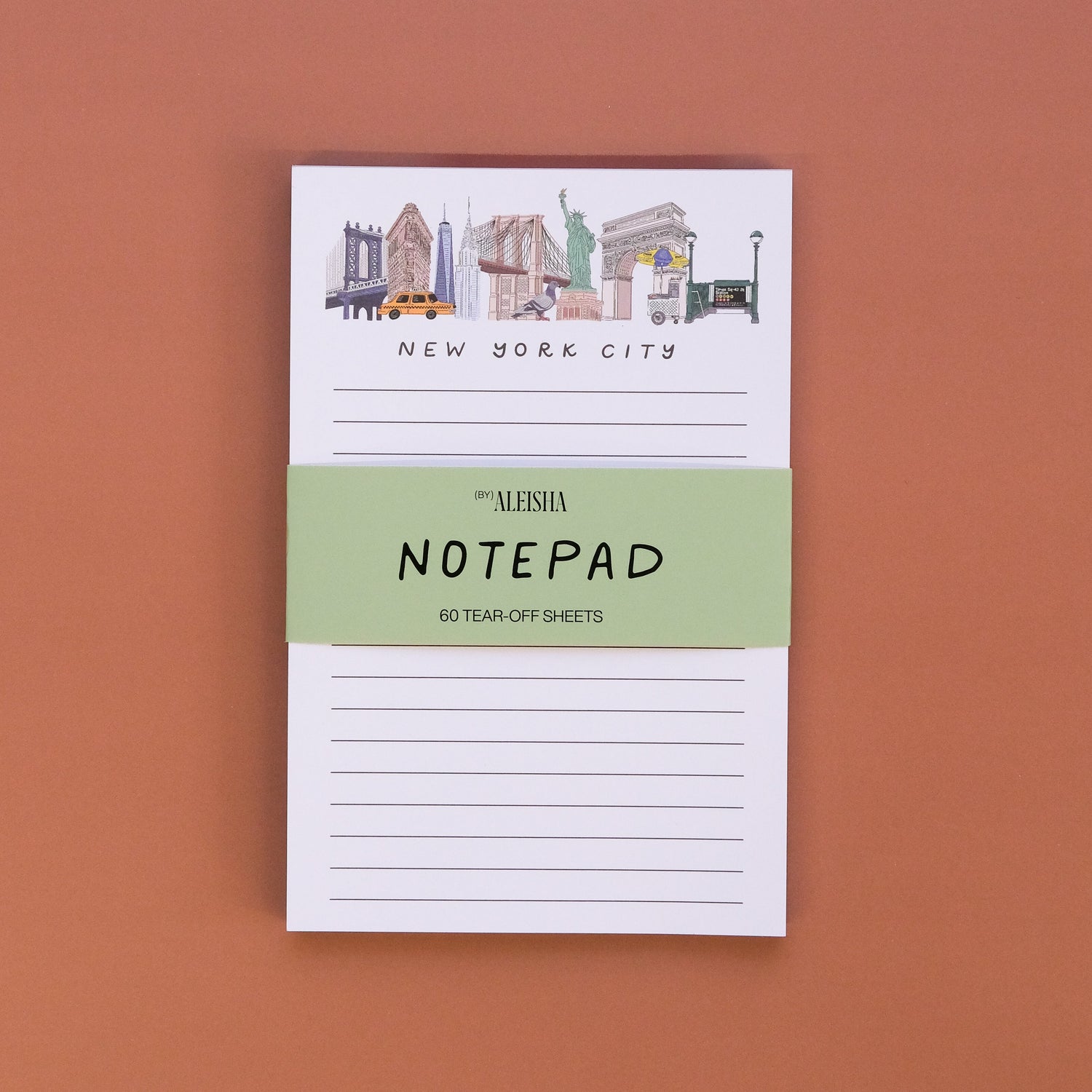 A small notepad with New York City skyline illustrations at the top, held by a light green paper band reading 'ALESHA NOTEPAD' on a solid terracotta background.